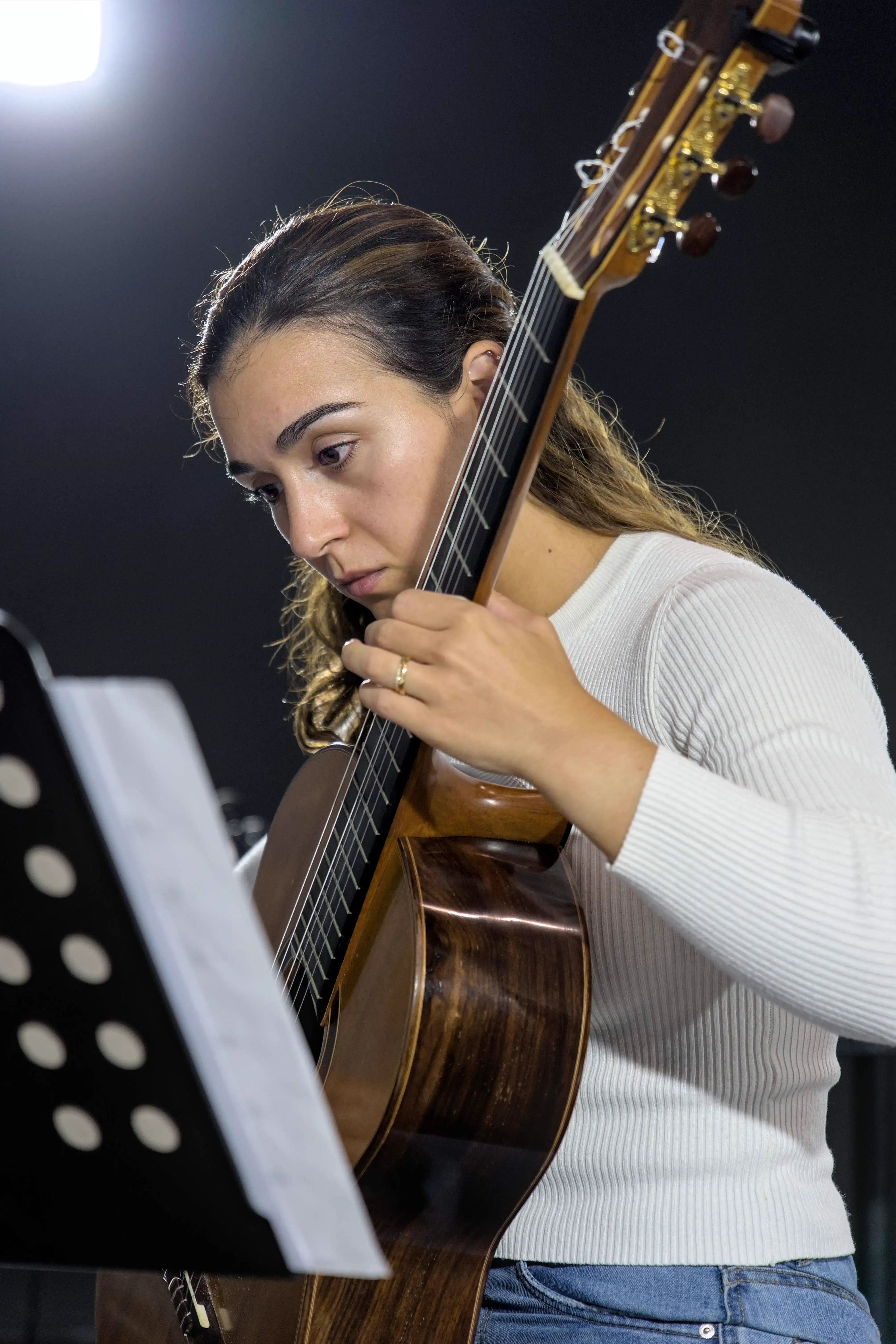 Uma mulher toca guitarra clássica enquanto olha a partitura. Ela tem cabelo claro apanhado e tem vestida uma camisola branca.